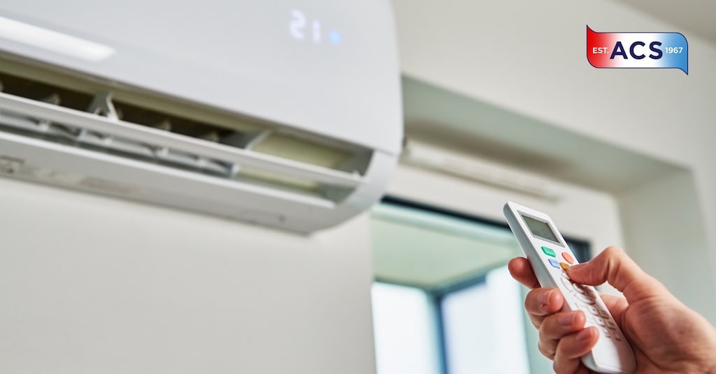 A person uses a remote control to adjust a wall-mounted air conditioner; the display shows 21°C. The ACS logo is visible in the top right corner.