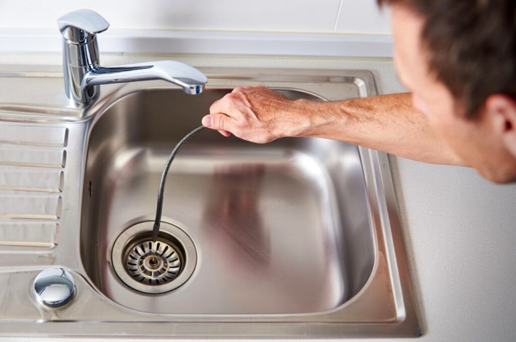 A person in Putnam County, GA uses a plumbing snake tool to unclog a stainless steel kitchen sink.