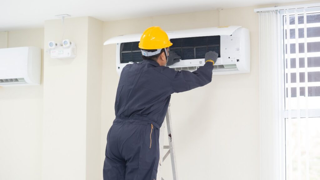 A worker in a helmet and coveralls stands on a ladder in Covington, inspecting or repairing a wall-mounted air conditioner indoors. Covington, GA