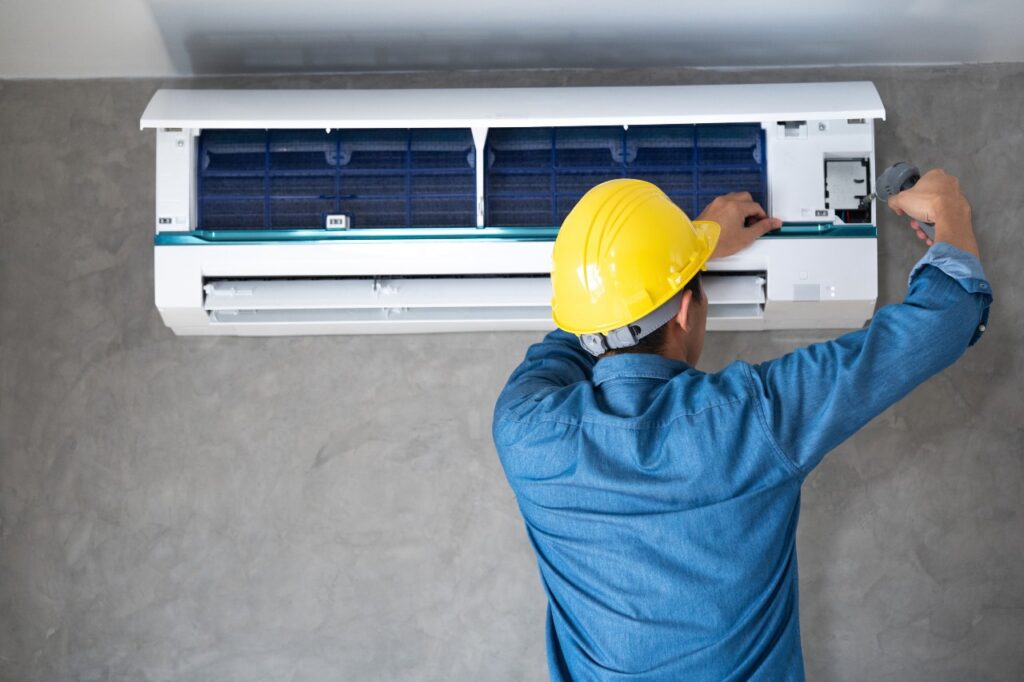 A worker in a yellow hard hat installs or repairs a wall-mounted air conditioner with a screwdriver in Dublin.