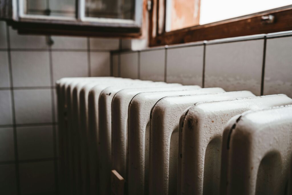 %Focuskw% | Air Conditioning Specialists A close-up view of a white, old-fashioned radiator beneath a partially open window in a tiled room.